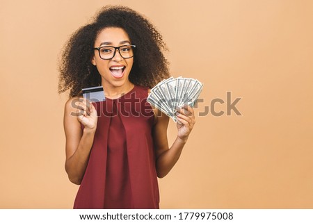 Happy winner. Portrait of african american successful woman 20s with afro hairstyle holding lots of money dollar banknotes and credit card isolated over beige background. 