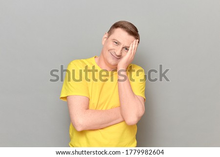 Studio portrait of happy cheerful blond mature man wearing bright yellow T-shirt, propping head with hand, smiling, laughing joyfully at funny jokes, standing isolated over gray background