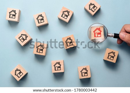 Hand holding magnifier glass to searching red house icon on wooden cube block among black house icon. Finding the best house and real estate for family concept.