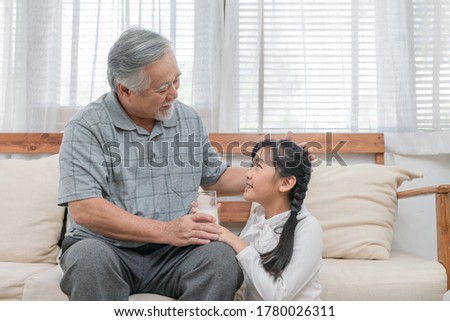 Asian granddaughter giving some milk to grandfather while sitting on sofa in the living room