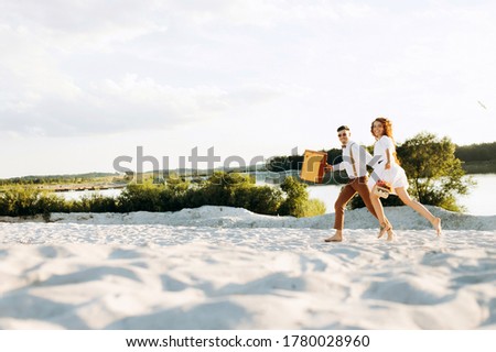 Happy couple running on the sand, sunset, stylish couple with a suitcase and flowers in a basket.