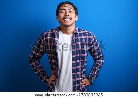 Asian student man smiled at the camera looking confident while putting both hands on the waist against blue background. Studio shots