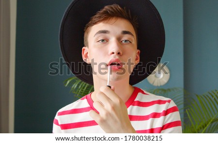 Young man holding lipstick or lip gloss and paints lips prepare getting ready looking in camera like mirror. Gay make up. Royalty-Free Stock Photo #1780038215