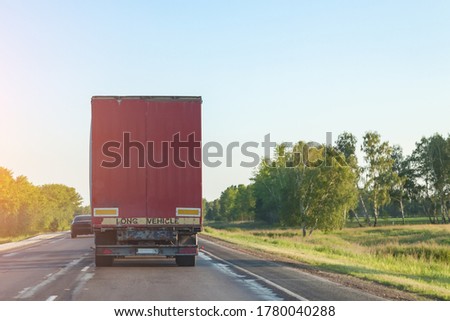 Rear view of a truck with a red body on a road, delivering and transporting cargo by a transport company over long distances through the countryside.