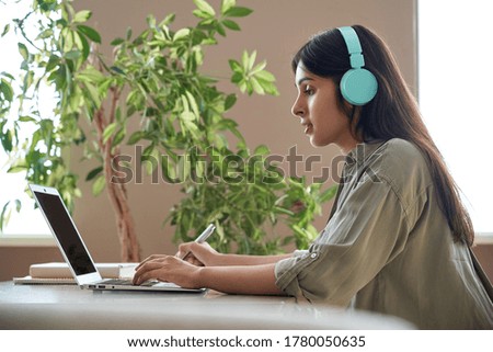 Indian teen girl school college student wear headphones learning watching online webinar class looking at laptop computer elearning lesson making notes or video calling virtual meeting remote teacher. Royalty-Free Stock Photo #1780050635