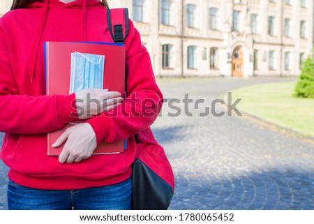 First day at school after university concept. Cropped close up photo of teen girl in casual red sweater holding books and medical mask standing near doors enter
