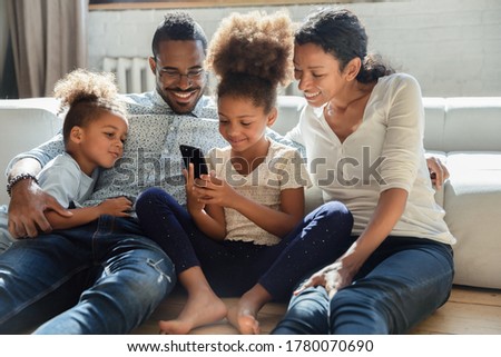 Smiling loving young biracial parents with little children sit relax on sofa in living room using cellphone, happy african american family with small kids watch video or play on smartphone together