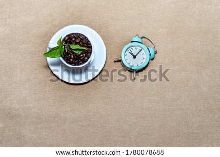 Roasted coffee beans In a white coffee cup has green leaves and an alarm clock and on a light brown fabric background and a few coffee beans scattered.  From top view