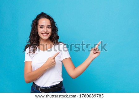young pretty woman smiling happily and pointing to side and upwards with both hands showing object in copy space against blue background