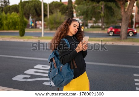 African American hipster girl dressed in trendy casual clothes with smartphone reading news on social media standing at bus stop, positive female tourist