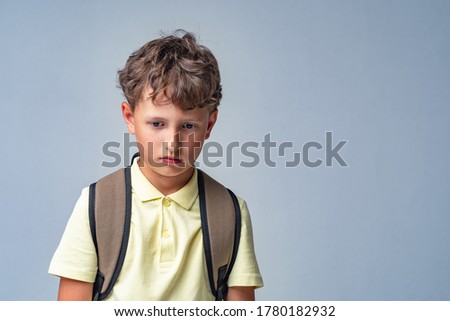 sad schoolboy with a backpack, standing on a gray background. the child is upset, problems with school, problems in communication, failure.