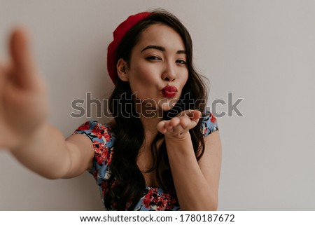 Pretty brunette curly woman in red beret and stylish floral blouse blows kiss, takes selfie and poses on white background.