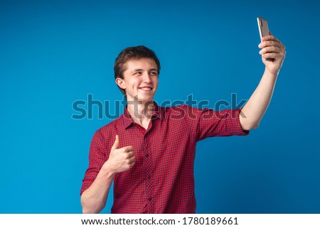 young cheerful man takes a selfie on his smartphone, dressed in a checked shirt. A handsome guy looks at his mobile phone takes photos and gives a thumbs up on a blue background.