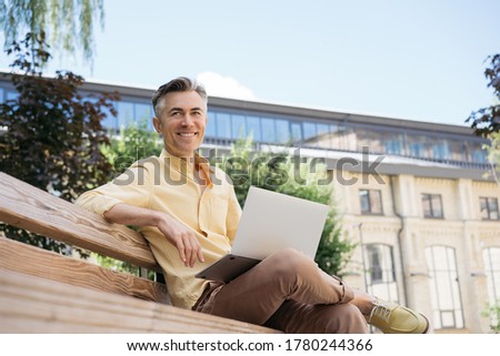 Handsome mature businessman using laptop computer, planning start up, sitting outdoors. Smiling freelancer working in park, sitting on bench. Successful business concept