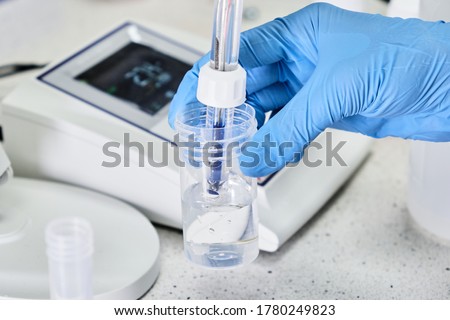 Woman in a rubber gloves holding a glass electrode for measuring of pH of the solution using pH meter. Analytical or electro chemistry laboratory. 