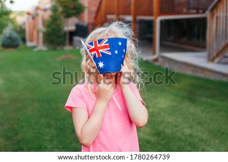 Adorable cute happy Caucasian girl holding Australian flag. Funny child kid covering her face with Australia flag. Little citizen celebrating Australia Day holiday in January outdoor.
