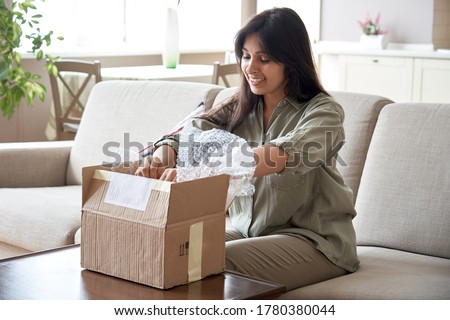 Smiling indian woman shopper customer opening post package box sitting on couch at home. Happy female consumer unpacking parcel receiving postal shipping delivery service order retail product purchase Royalty-Free Stock Photo #1780380044