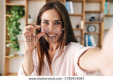 Image of joyful adult businesswoman smiling and holding eyeglasses while taking selfie photo in office