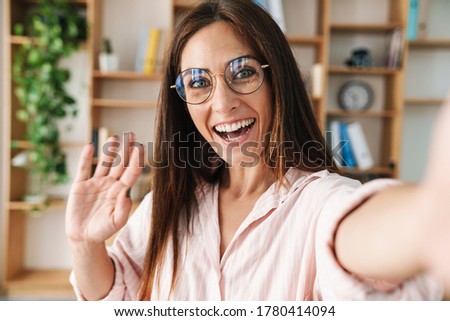 Image of excited adult businesswoman waving hand while taking selfie photo in office