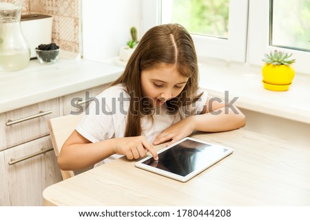 Little girl at home in the kitchen sitting at the table with tablet looking at device screen watching cartoons enjoy, preschool modern tech user parental control safety, gadget overuse concept