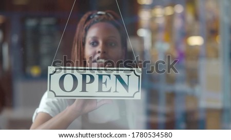 Small business african female owner smiling while turning sign for opening of cafe. Happy afro-american waitress in apron turning sign on door opening restaurant in morning.