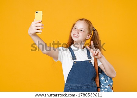Young school girl 12-13 years old in white t-shirt backpack hold in hand using mobile cell phone doing selfie isolated on yellow background children studio portrait Kids education lifestyle concept.