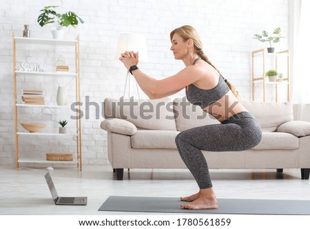 Quarantine and exercise at home. Slim adult woman in sport uniform with fitness tracker crouches and watches at laptop in living room interior, copy space Royalty-Free Stock Photo #1780561859