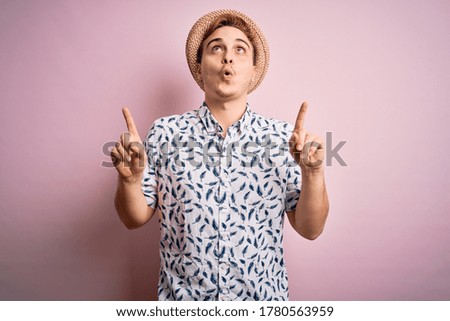 Handsome redhead man on vacation wearing casual shirt and hat over isolated pink background amazed and surprised looking up and pointing with fingers and raised arms.