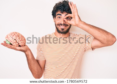 Handsome young man with curly hair and bear holding brain as mental health concept smiling happy doing ok sign with hand on eye looking through fingers 