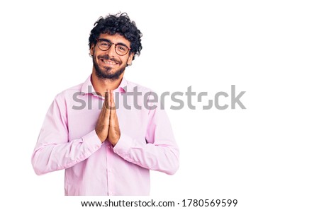 Handsome young man with curly hair and bear wearing business clothes praying with hands together asking for forgiveness smiling confident. 