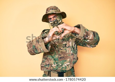 Young handsome man wearing camouflage army uniform and balaclava smiling in love showing heart symbol and shape with hands. romantic concept. 