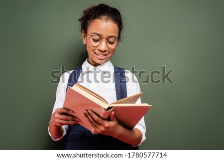 smiling african american schoolgirl reading book near empty green chalkboard