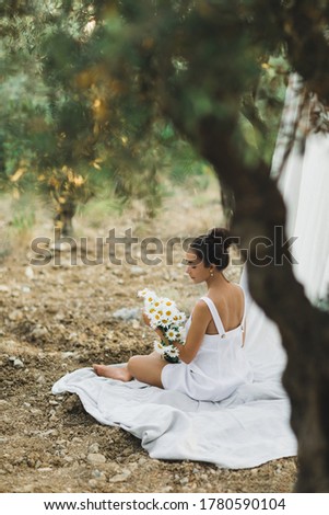 Portrait of young brunette woman in white summer linen dress on background of hanging textile cloth. Relaxation and leisure, picnic in olive garden, bouquet of chamomile flowers.
