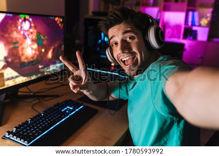 Image of cheerful man gesturing peace sign and taking selfie photo while playing video game on computer at home