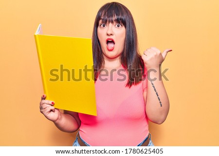 Young plus size woman holding book pointing thumb up to the side smiling happy with open mouth 