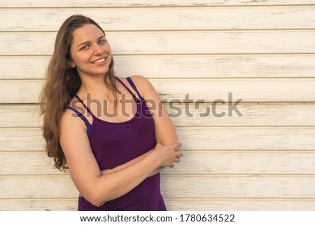 Joyful young woman with long blonde and arms crossed hair standing next to a white wood wall
