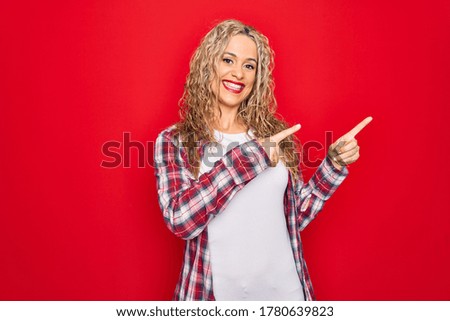 Young beautiful blonde woman wearing casual shirt standing over isolated red background smiling and looking at the camera pointing with two hands and fingers to the side.