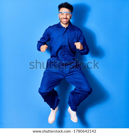 Young handsome hispanic man wearing painter uniform and glasses smiling happy. Jumping with smile on face doig winner sign with fists up over isolated blue background