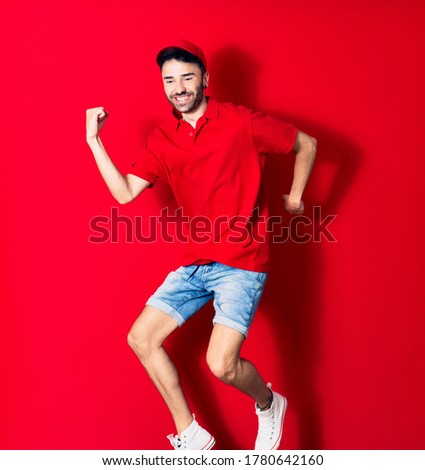Young handsome deliveryman wearing cap smiling happy. Jumping with smile on face over isolated red background