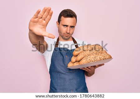 Young handsome baker man with blue eyes wearing apron holding tray with wholmeal bread with open hand doing stop sign with serious and confident expression, defense gesture