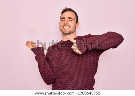 Young handsome man with blue eyes wearing casual sweater standing over pink background Showing palm hand and doing ok gesture with thumbs up, smiling happy and cheerful