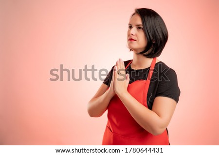 Woman employed at supermarket with red apron and black t-shirt, praying isolated on red background