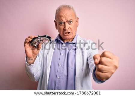 Middle age hoary optical man wearing coat holding optometry glasses over pink background annoyed and frustrated shouting with anger, crazy and yelling with raised hand, anger concept