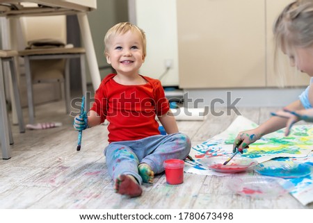 Adorable cute caucasian little blond siblings children enjoy having fun painting with brush and palm at home indoors . Cheerful happy kids smiling drawing masterpiece art picture. Messy dirty room