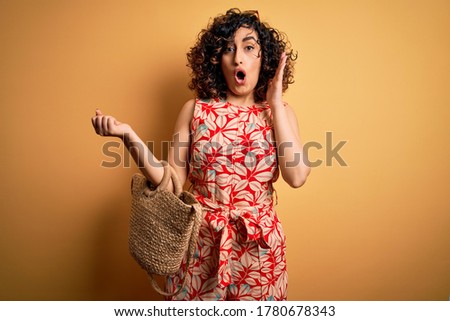Young curly arab woman on vacation wearing floral dress and sunglasses holding wicker bag scared in shock with a surprise face, afraid and excited with fear expression