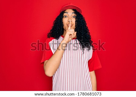 Young african american curly sportswoman wearing baseball cap and striped t-shirt asking to be quiet with finger on lips. Silence and secret concept.