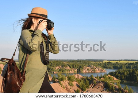 Woman in a long green shirt and yellow hat makes a landscape photo. She holds a leather backpack. Mountains and a lake on background. Evening light. Clear blue sky