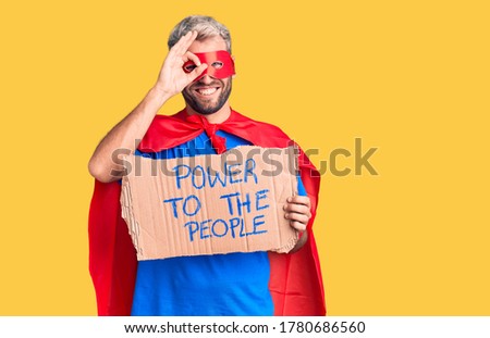 Young blond man wearing super hero custome holding power to the people cardboard banner smiling happy doing ok sign with hand on eye looking through fingers 