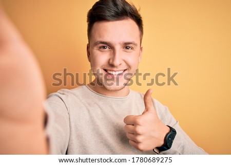 Young handsome caucasian man talking a selfie picture over yellow isolated background happy with big smile doing ok sign, thumb up with fingers, excellent sign