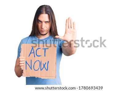 Young adult man with long hair holding act now banner with open hand doing stop sign with serious and confident expression, defense gesture 